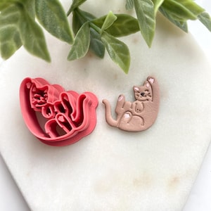 May include: A pink cat-shaped cookie cutter and a tan cat-shaped cookie. The cutter is made of plastic and has a detailed design. The baked cookie is lying on its back with its paws up. The cat cookie has pink accents.