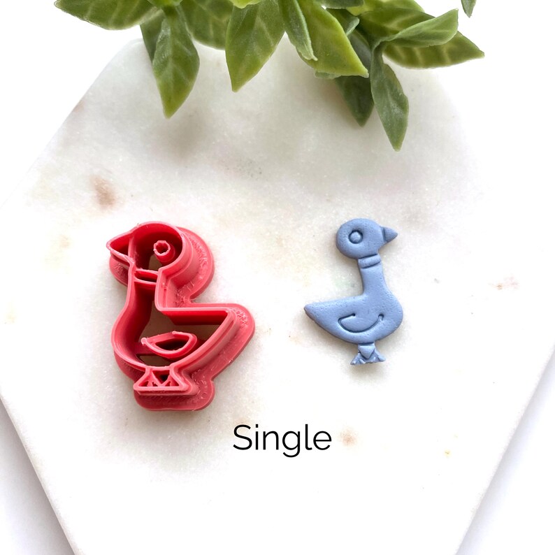 May include: A pink plastic bird-shaped cookie cutter next to a light blue bird-shaped cutout. The word Single is printed below the objects. The cutter features a detailed outline of a bird with a circular eye, a wing, and feet, matching the cutout.