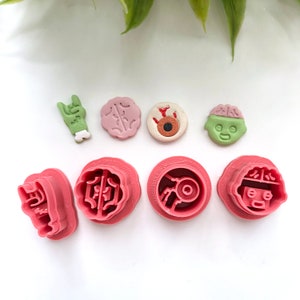 May include: A selection of Halloween-themed cookie cutters and decorated biscuits. The cutters are pink and shaped like a skull, brain, and eyeball. The biscuits are decorated with green, pink, and brown icing, depicting a zombie hand, brain, and eyeball.