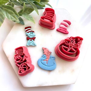 Peut inclure: Ensemble de découpoirs à biscuits en plastique rouge et rose, représentant un chapeau, un bocal à poissons et un personnage bleu avec un nœud papillon rouge. Les découpoirs sont disposés sur une surface en marbre blanc.