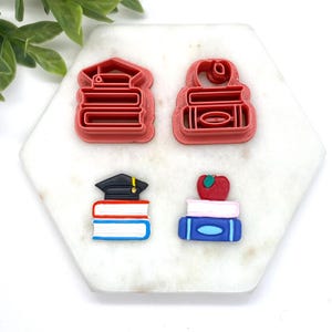 May include: Four red and blue silicone cookie cutters shaped like stacks of books. Two of the cutters have a graduation cap on top of the books, and one has an apple on top. The cutters are on a white background.