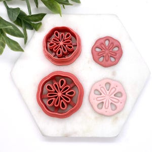 May include: Four cookie cutters, two red and two pink, with a flower design. The cutters are on a white marble surface.