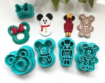 Könnte beinhalten: Ein Set aus türkisfarbenen Kunststoff-Ausstechformen und passenden Fondant-Auflegern. Die Formen umfassen eine Schleife, einen Kranz, einen Schneemann, einen Nussknacker und einen Lebkuchenmann, alle mit charakteristischen Mausohren.