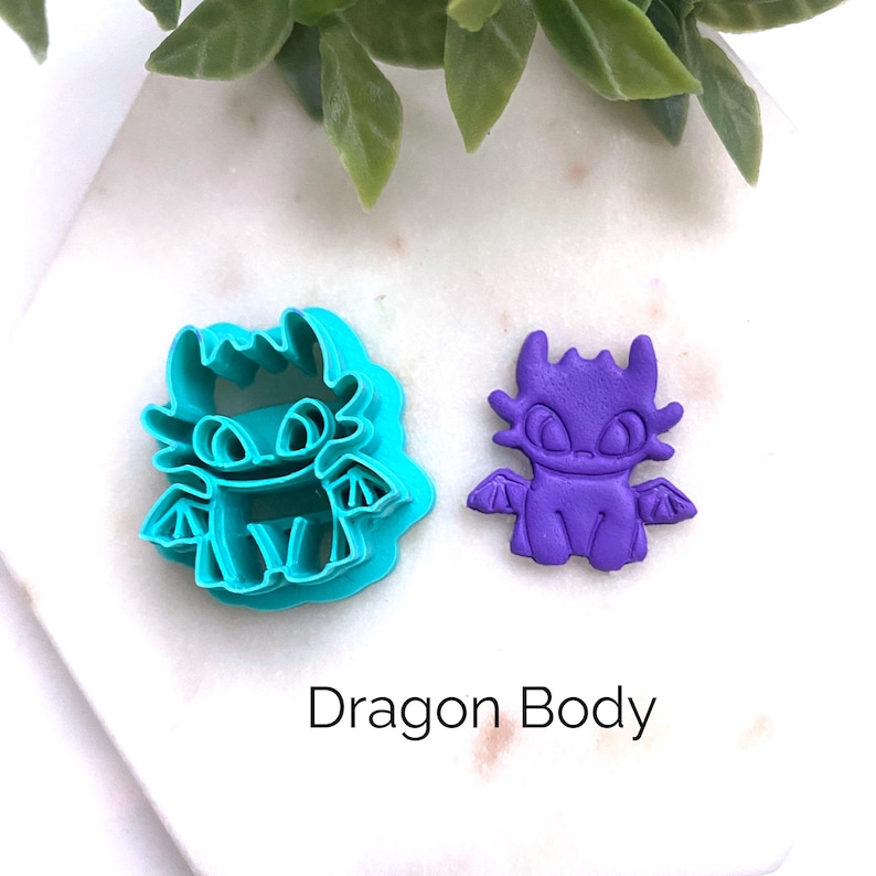 K&ouml;nnte beinhalten: Ein t&uuml;rkisfarbener Ausstechform aus Kunststoff in Drachenform und eine passende lilafarbene Drachenfigur aus Ton. Das Drachendesign zeigt gro&szlig;e Augen, spitze Ohren und kleine Fl&uuml;gel. Der Text "Dragon Body" ist darunter in Schwarz gedruckt.