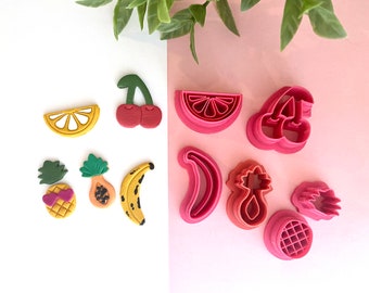 May include: A set of nine colorful plastic cookie cutters shaped like various fruits, including a pineapple, banana, lemon, watermelon, and cherries. The cutters are on a pink and white background.