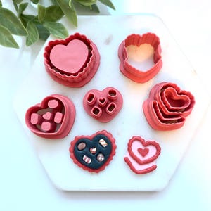 May include: A collection of pink heart-shaped cookie cutters in various designs, including layered hearts and a heart-shaped candy box. The cutters are arranged on a white hexagonal surface, perfect for Valentine's Day baking.