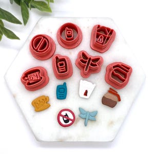 May include: A set of 12 red, blue and brown plastic cookie cutters shaped like a dragonfly, a phone, a jar, a takeaway container, a house, a stop sign, a doughnut, a coffee cup, a 'take away' sign, a 'likes' sign and a 'ticket' sign. The cutters are arranged on a white marble surface.