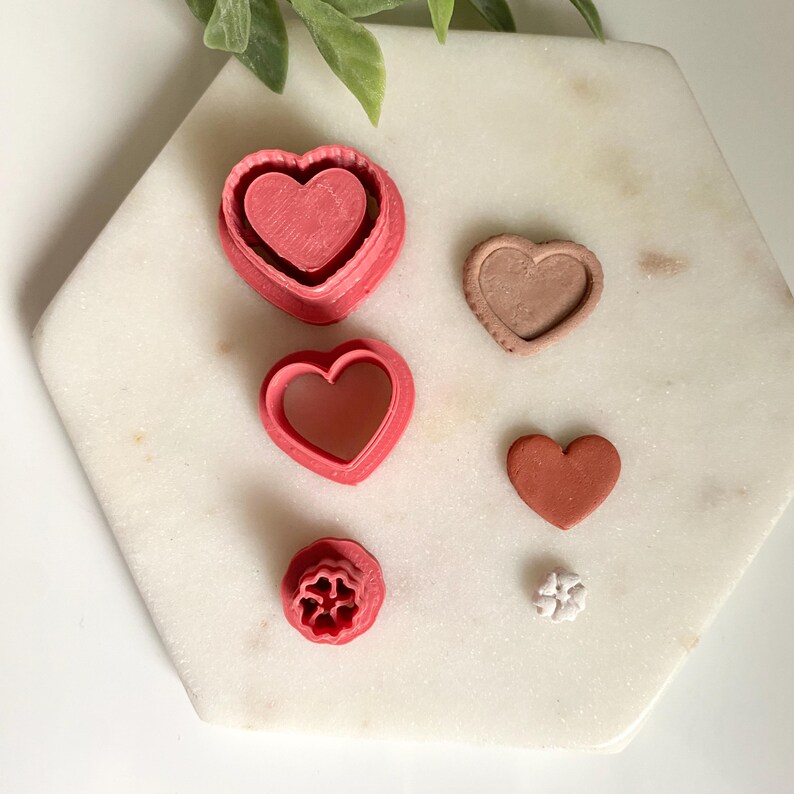 Op de afbeelding: Een verzameling roze en bruine hartvormige koekjessnijders en een klein hartvormig koekje op een wit marmeren oppervlak. De snijders vari&euml;ren in grootte en ontwerp, waaronder een genest hart en een bloemvorm. Een takje groene bladeren op de achtergrond.