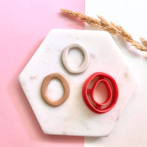 May include: Three clay circles, one beige, one white, and one red, on a white marble hexagonal tile. A red plastic cookie cutter with a double-ring design is also on the tile.