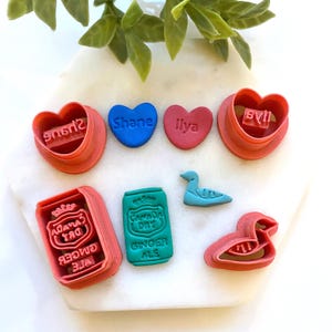 May include: A selection of colourful cookie cutters and shaped treats. Includes heart-shaped cutters with names, a Canada Dry ginger ale can, and a duck. The colours are red, blue, and teal.