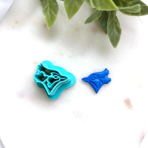 May include: A turquoise plastic cookie cutter shaped like a blue jay bird profile, accompanied by a smaller, solid blue plastic cutout of the same bird shape, featuring detailed outlines of the head, beak, and wing feathers in a flat design.