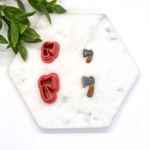 May include: A collection of miniature axe-shaped items. Two pink plastic cookie cutters are next to two small axe-shaped earrings. The earrings have gray axe heads and brown wooden handles. The items are arranged on a white hexagonal surface.