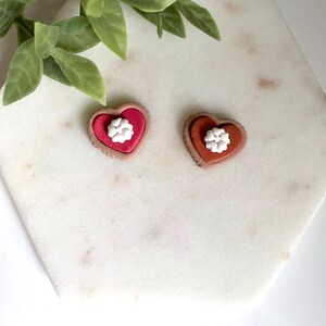 Op de afbeelding: Twee hartvormige oorbellen. E&eacute;n is rood met een witte bloem, de andere is oranje met een witte bloem. De oorbellen hebben een bruine rand. De oorbellen liggen op een wit marmeren oppervlak met groene bladeren op de achtergrond.
