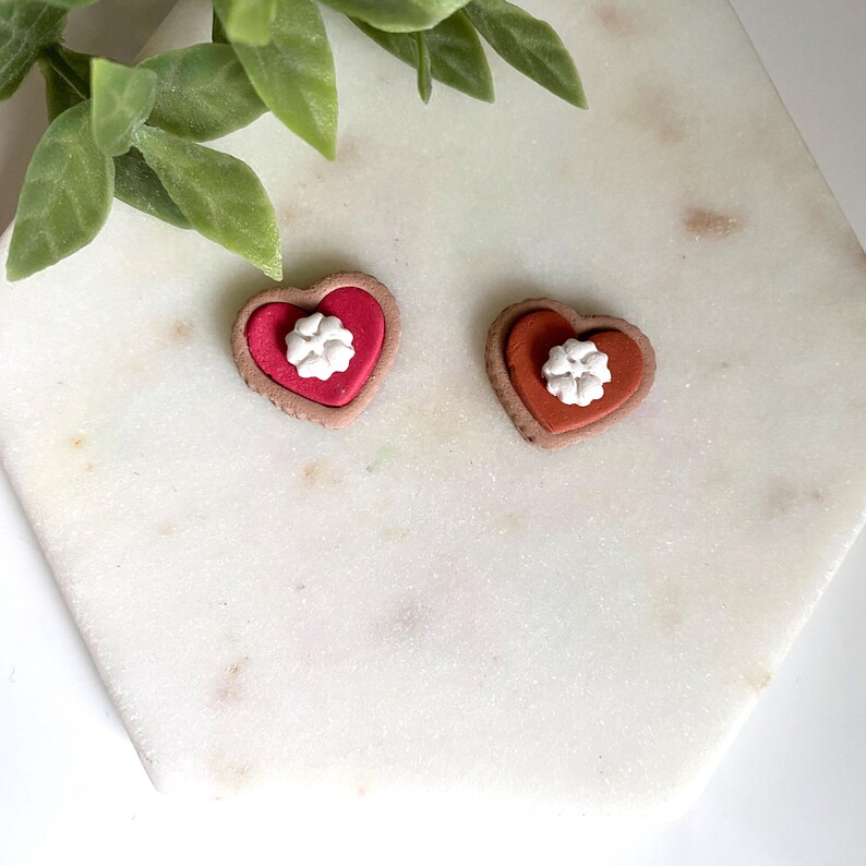 Op de afbeelding: Twee hartvormige oorbellen. E&eacute;n is rood met een witte bloem, de andere is oranje met een witte bloem. De oorbellen hebben een bruine rand. De oorbellen liggen op een wit marmeren oppervlak met groene bladeren op de achtergrond.