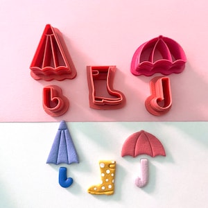 May include: Pink plastic cookie cutters in the shapes of an umbrella, rain boots, and a raindrop. The umbrella and raindrop are also in blue and the rain boots are yellow with white polka dots. The cutters are arranged on a pink and white background.