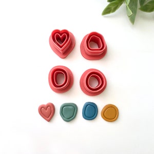 May include: A collection of pink polymer clay cutters in various organic shapes, including a heart and rounded forms. Below are finished clay pieces in coordinating colors: dusty rose, sage green, navy blue, and mustard yellow. Ideal for crafting.
