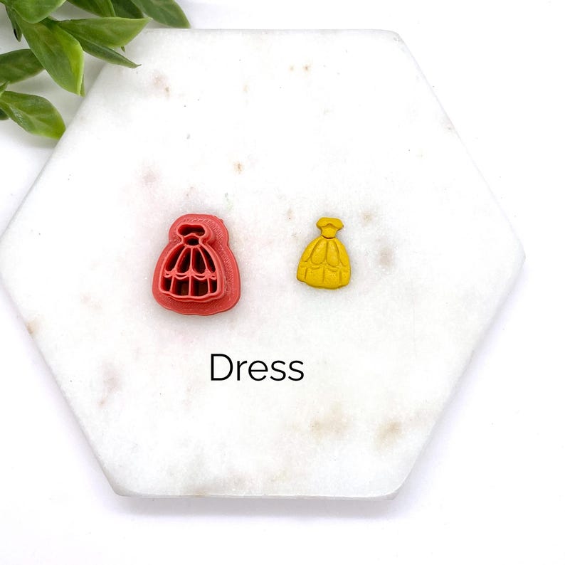 Peut inclure: Deux petits emporte-pi&egrave;ces en forme de robe, l'un rouge et l'autre jaune, sur une surface en marbre blanc. Le texte "Dress" est imprim&eacute; sous les emporte-pi&egrave;ces.