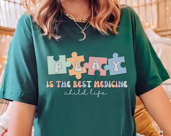 Op de afbeelding: Een groen T-shirt met een kleurrijk puzzelstukontwerp met de tekst "PLAY IS THE BEST MEDICINE child life".