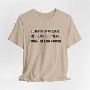 May include: A beige t-shirt with the text "I'D RATHER BE LOST IN COLUMBUS THAN FOUND IN ANN ARBOR" printed in black. The shirt is made of a soft material and has short sleeves.