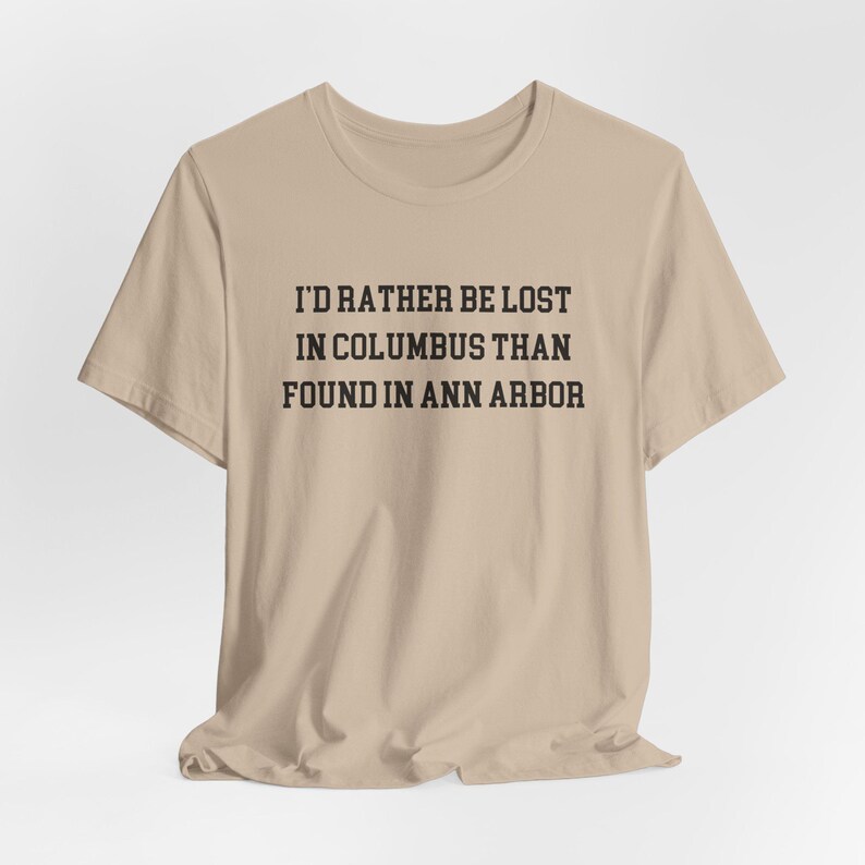 May include: A beige t-shirt with the text "I'D RATHER BE LOST IN COLUMBUS THAN FOUND IN ANN ARBOR" printed in black. The shirt is made of a soft material and has short sleeves.