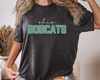 May include: Dark gray t-shirt with the text "ohio BOBCATS" in white and green. The word "ohio" is in a cursive font above the word "BOBCATS". The shirt is made of a soft material.