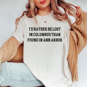 May include: Cream-colored t-shirt with the text "I'D RATHER BE LOST IN COLUMBUS THAN FOUND IN ANN ARBOR" in black. The shirt is worn by a person with long blonde hair and a gold necklace.