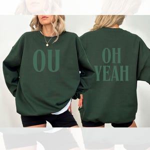Puede incluir: Sudadera verde oscuro con las letras "OU" estampadas en la parte delantera y "OH YEAH" en la trasera, ambas en verde a juego. La sudadera informal está diseñada para el uso diario.