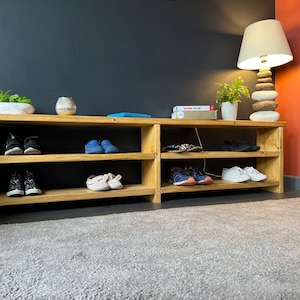 May include: A wooden shoe rack with three shelves, filled with shoes. The rack is in a hallway with a gray wall, a rug, and a lamp with a white shade.