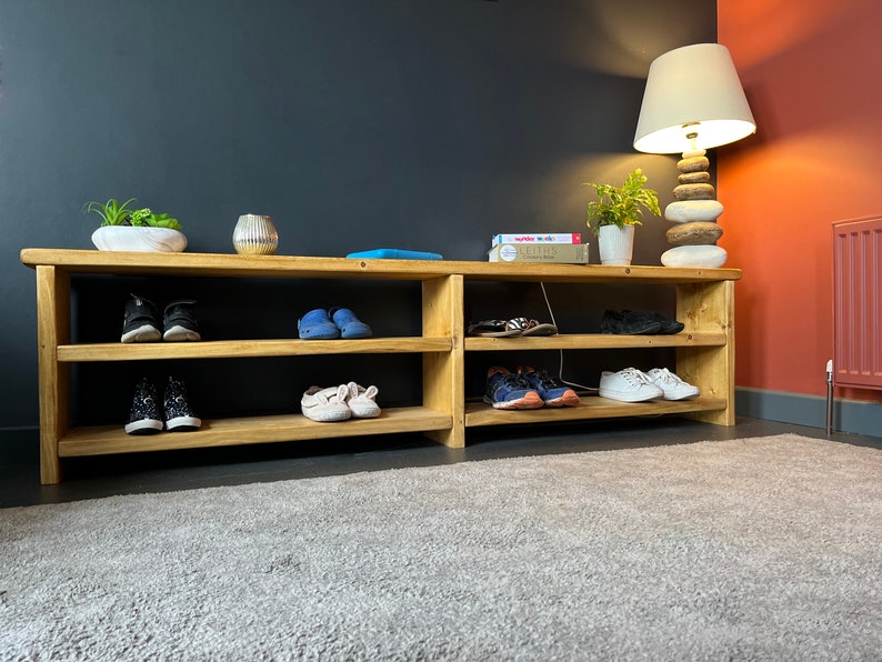 May include: A wooden shoe rack with three shelves, filled with shoes. The rack is in a hallway with a gray wall, a rug, and a lamp with a white shade.