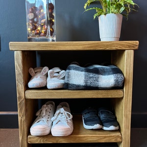 May include: A wooden shoe rack with three shelves. The top shelf has a black and white plaid blanket, the middle shelf has two pairs of white sneakers, and the bottom shelf has two pairs of black sneakers. There is a glass vase with chestnuts and lights on the top of the rack.