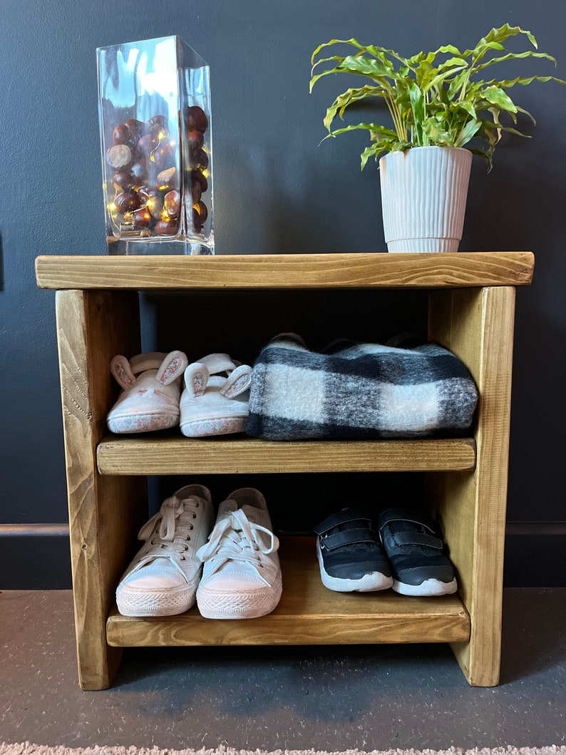 May include: A wooden shoe rack with three shelves. The top shelf has a black and white plaid blanket, the middle shelf has two pairs of white sneakers, and the bottom shelf has two pairs of black sneakers. There is a glass vase with chestnuts and lights on the top of the rack.