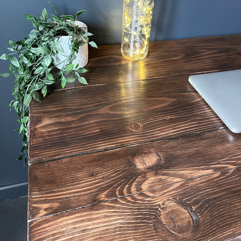 K&ouml;nnte beinhalten: Ein dunkelbrauner Holztisch mit nat&uuml;rlicher Holzmaserung. Der Tisch hat eine hellbraune Pflanze in einem wei&szlig;en Topf auf der linken Seite und einen Laptop auf der rechten Seite.