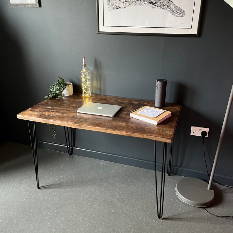 K&ouml;nnte beinhalten: Ein dunkler Holztisch mit Haarnadelbeinen. Der Tisch hat einen Laptop, ein Notizbuch und eine Glasflasche mit Lichtern darauf. Der Tisch steht vor einer dunklen Wand.