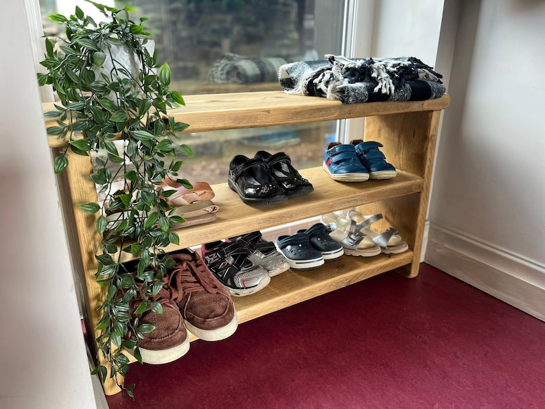 May include: A wooden shoe rack with three shelves, holding a variety of shoes, including sneakers, sandals, and dress shoes. A green plant hangs from the top shelf.