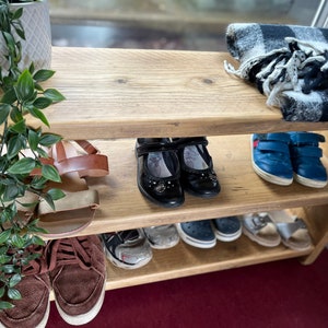 May include: A wooden shoe rack with three shelves. The top shelf has a black and white plaid throw blanket. The middle shelf has a pair of brown sandals, a pair of black flats, and a pair of blue sneakers. The bottom shelf has a pair of brown sneakers, a pair of black sandals, and a pair of blue sandals.