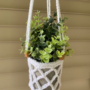 May include: White crocheted hanging planter with a green plant inside. The planter has wooden beads on the top of the cords.