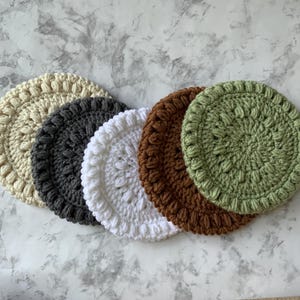 May include: Five crocheted coasters in different colors and patterns. The coasters are round and have a raised texture. The colors are beige, gray, white, brown, and green.