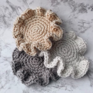 Op de afbeelding: Drie gehaakte onderzetters in beige, grijs en wit met een gekrulde rand.