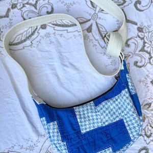 May include: A blue and white houndstooth pattern shoulder bag with a white strap. The bag features a curved shape and is made of quilted fabric. The strap is adjustable and made of a sturdy, off-white material.