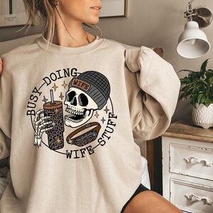 May include: A beige sweatshirt with a graphic print of a skull wearing a beanie with the word "Wife" on it. The skull is holding a coffee cup with a leopard print design and a fanny pack. The text around the skull reads "Busy Doing Wife Stuff".