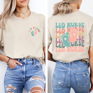 May include: A beige t-shirt with a graphic of a pair of baby footprints and the text "L&D NURSE" on the front. The back of the shirt has the text "L&D NURSE" repeated in a rainbow pattern with a heart in the middle of each graphic.