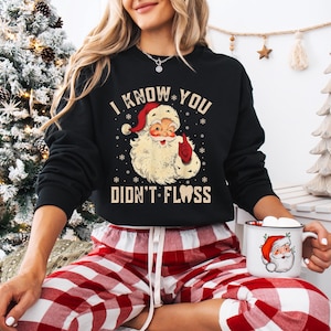 May include: Black sweatshirt with the text "I KNOW YOU DIDN'T FLOSS" and a Santa Claus graphic. The Santa graphic has a red hat and mitten. The person is wearing red and white plaid pajama pants and holding a mug with a Santa Claus graphic.