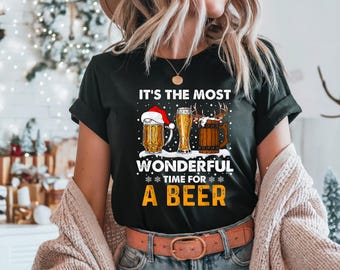 Może przedstawiać: Czarny t-shirt z napisem "IT'S THE MOST WONDERFUL TIME FOR A BEER". Grafika przedstawia trzy szklanki piwa, jedną z czapką Świętego Mikołaja i drewniany kufel z rogami. Koszulka jest noszona przez osobę.