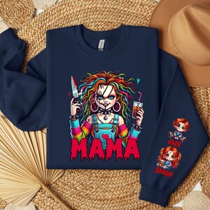 May include: Navy blue sweatshirt featuring a colorful graphic of a doll-like character holding a bloody knife and a drink. The word "MAMA" is printed in red. The sleeve has illustrations of two dolls with the names "Jack" and "Sarah".