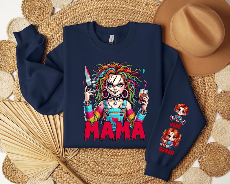 May include: Navy blue sweatshirt featuring a colorful graphic of a doll-like character holding a bloody knife and a drink. The word "MAMA" is printed in red. The sleeve has illustrations of two dolls with the names "Jack" and "Sarah".