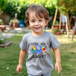 May include: A young child wearing a gray t-shirt with a blue car graphic and the text "rollin' into headstart".