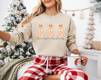 Może przedstawiać: Beżowa bluza z trzema pomarańczowymi kwiatowymi wzorami choinek i napisem "Tennessee Christmas" czarnym pismem. Osoba ma na sobie czerwono-białą piżamę w kratę i trzyma biały kubek z grafiką Świętego Mikołaja.
