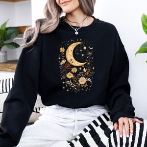 May include: Black sweatshirt with a crescent moon and floral design. The moon is gold and the flowers are a mix of yellow, orange, and brown.