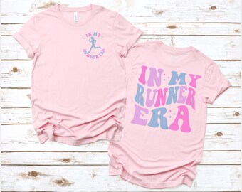 Könnte beinhalten: Pinkes T-Shirt mit dem Text "IN MY RUNNER ERA" in blauer, rosa und lila Schrift mit Sternen auf der Rückseite. Die Vorderseite des Shirts zeigt eine Silhouette eines Läufers mit dem gleichen Text in kleinerer Schrift.