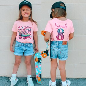 May include: A young person wearing a pink t-shirt with a roller skate graphic and the text "Rolling into 8". The person is also wearing denim shorts and a black baseball cap. A skateboard is held in the person's hand.